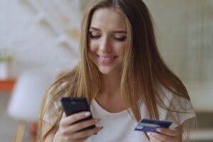 Wikypost: mujer joven revisando su crédito en el celular con su tarjeta en la mano