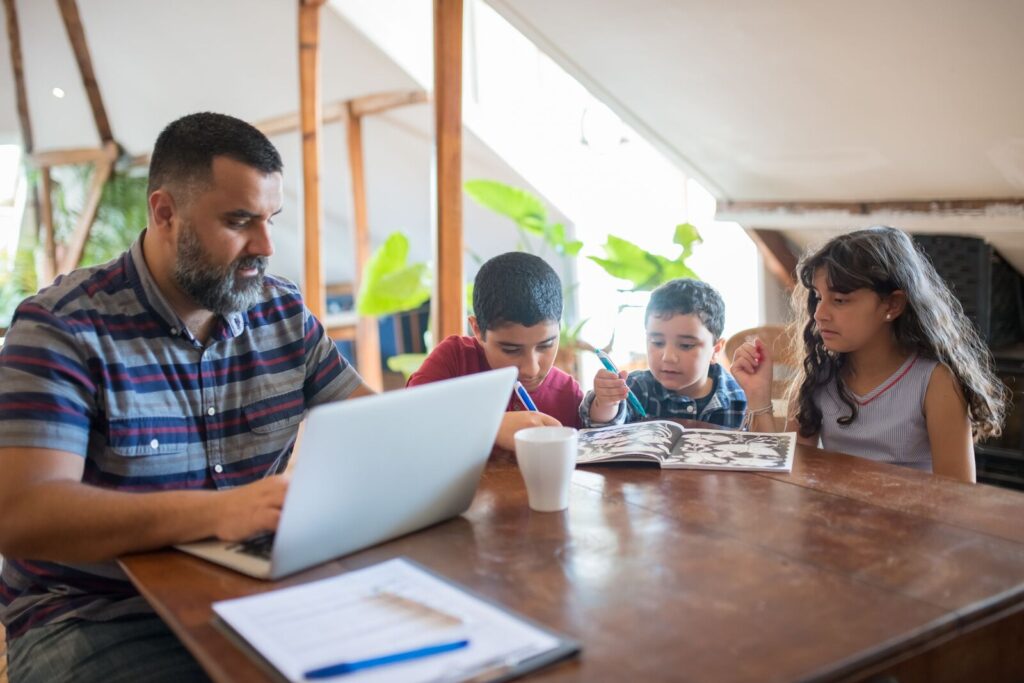 Presupuesto de una familia latina: padre con laptop y papeles mientras sus hijos dibujan en la mesa de la cocina